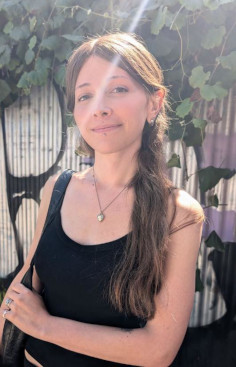 A smiling white woman with long hair, wearing a black tank top, standing outside on a sunny day.